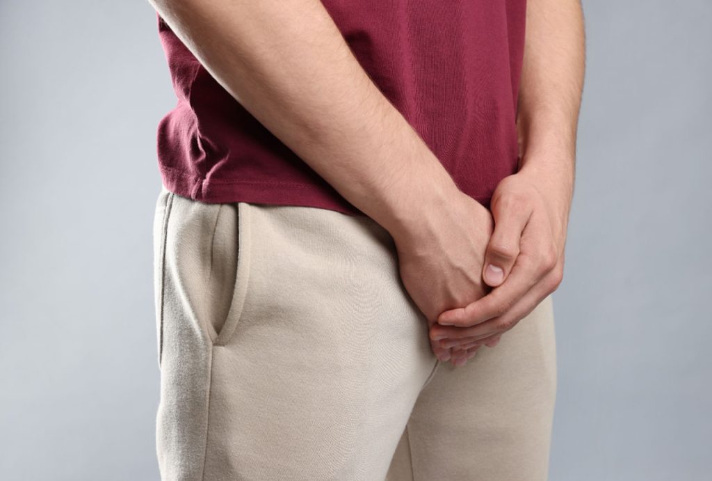 A man in beige sweatpants and a maroon shirt holds his groin area in clear discomfort, needing a gay men STD test in Houston.